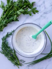 ranch dressing in a glass bowl with herbs surrounding it on a marble table