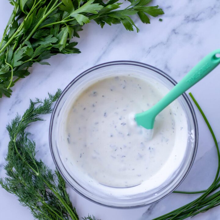 ranch dressing in a glass bowl with herbs surrounding it on a marble table