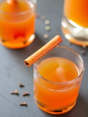 Apple cider in a clear glass on a black background with a cinnamon stick on top.