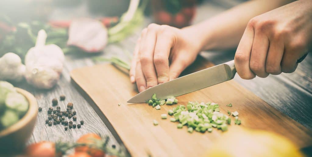 a knife cutting green onions on a cutting board surrounded by vegetables 