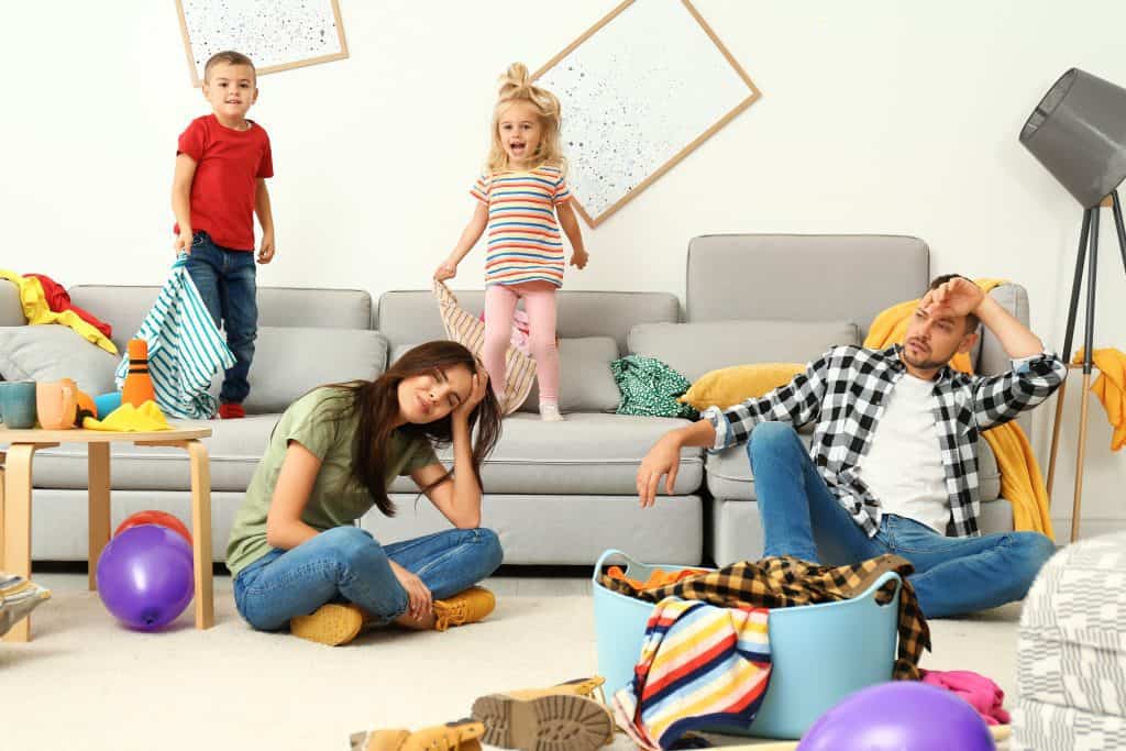 exhausted parents sitting on the floor in front of the couch while kids jump on it in a messy room