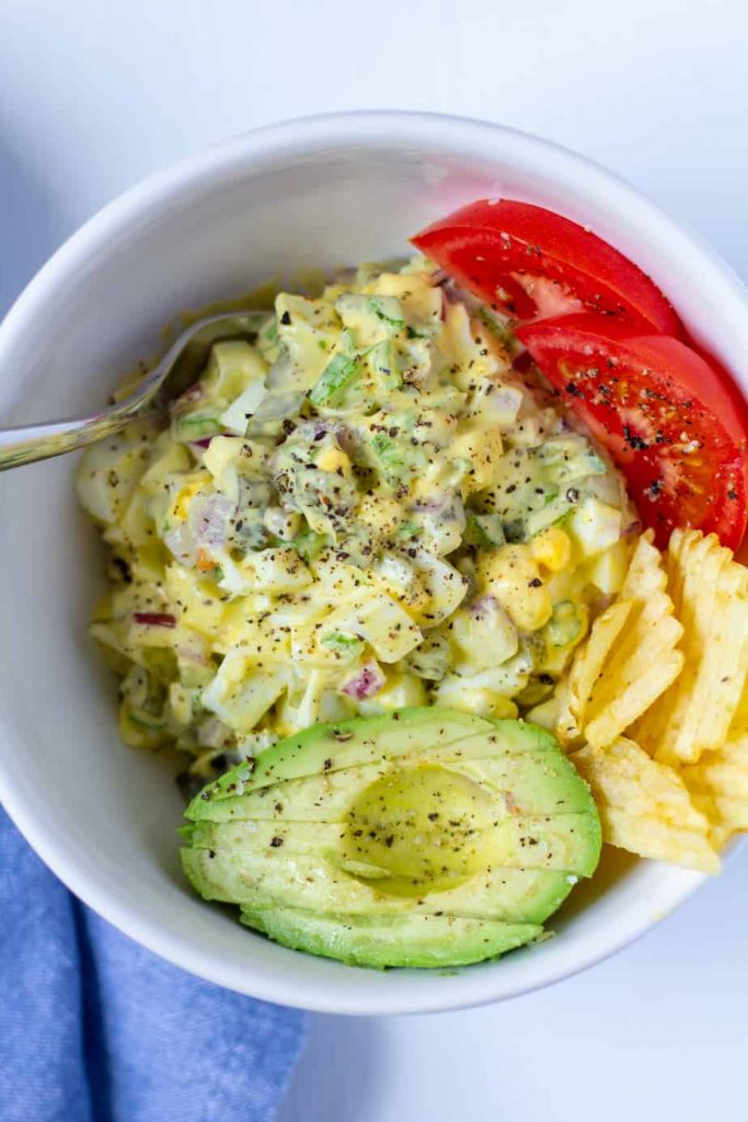 egg salad in a white bowl with avocados, potato chips, and tomato