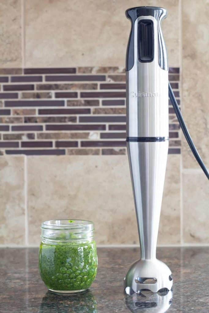 an immersion blender standing on a counter and sitting next to pesto