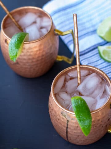 moscow mules in copper mugs on a black background with a white and blue striped napkin and sliced limes