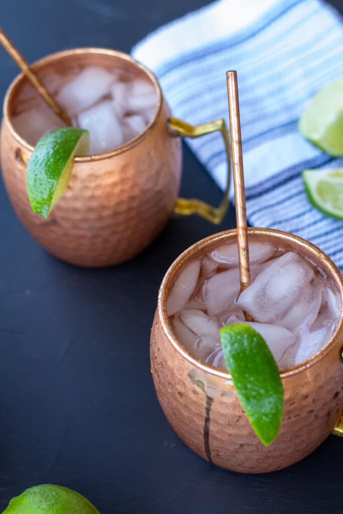 moscow mules in copper mugs on a black background with a white and blue striped napkin and sliced limes