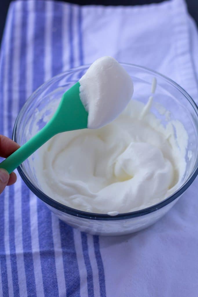 hard peaks whipped cream in a glass bowl with a green spoon on a white and blue background