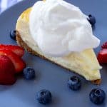 Whipped cream on a piece of cheesecake sitting on a blue plate with berries surrounding it