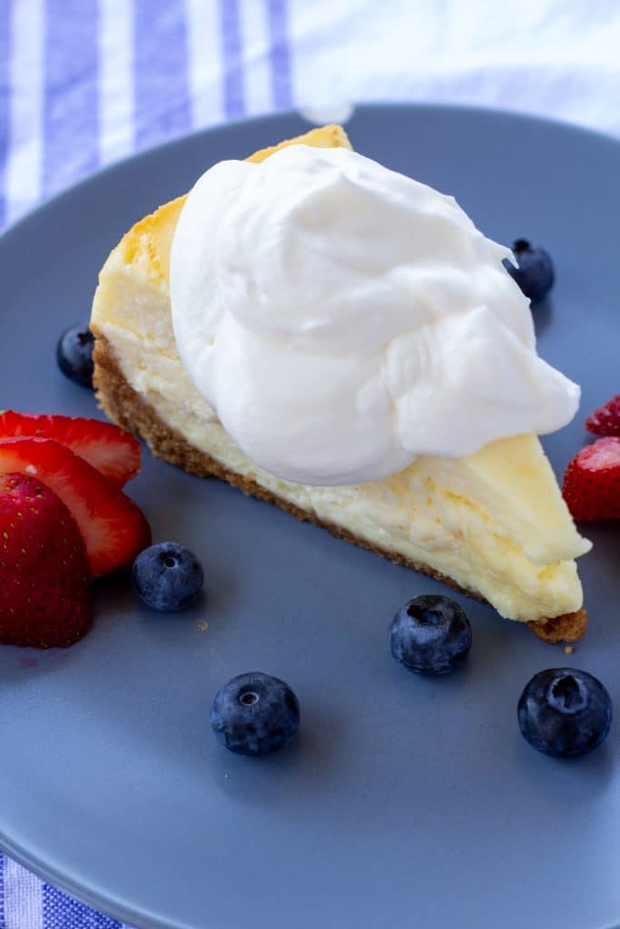 Whipped cream on a piece of cheesecake sitting on a blue plate with berries surrounding it