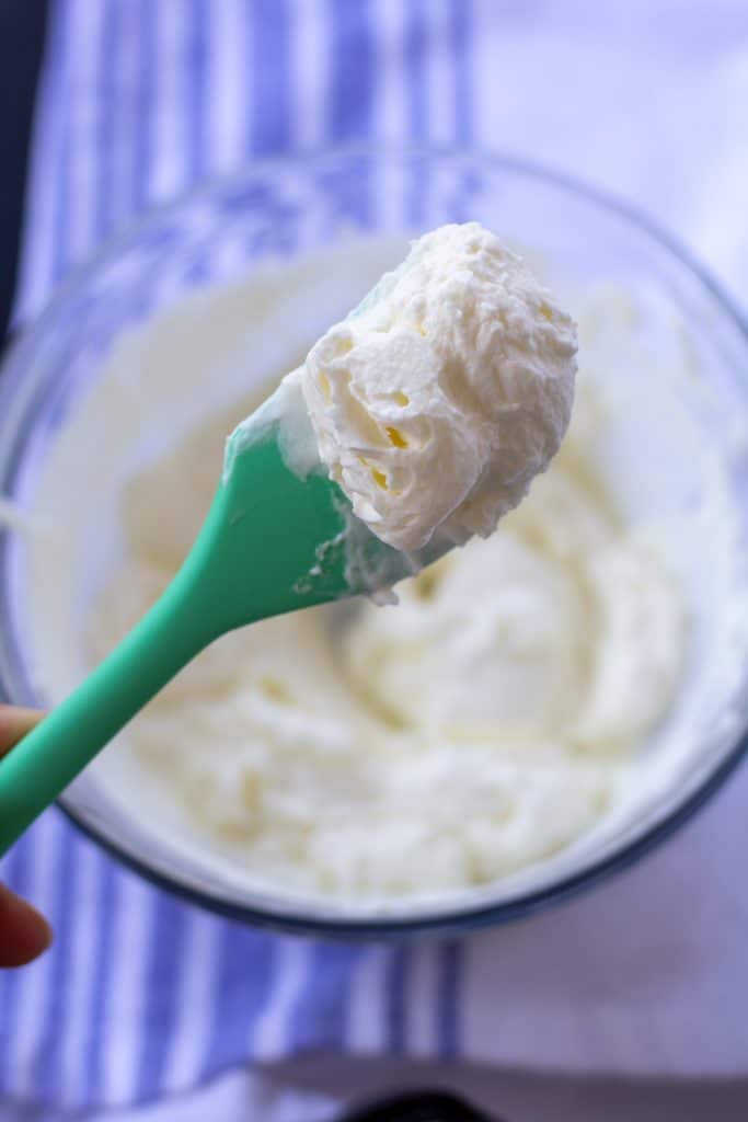 over whipped peaks whipped cream in a glass bowl with a green spoon on a white and blue background