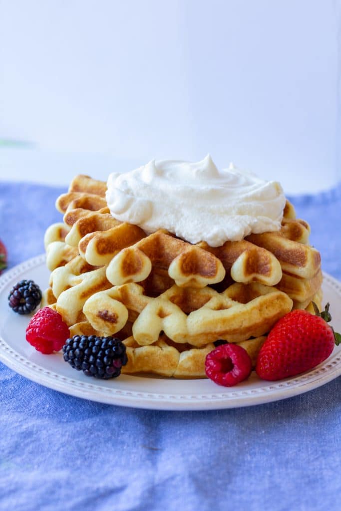 Breakfast waffles topped with whipped cream surrounded by berries on a white plate with a blue and white background. 