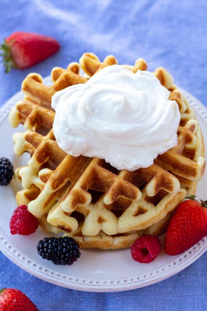 a stack of waffles with whipped cream, on a white plate, surrounded by berries