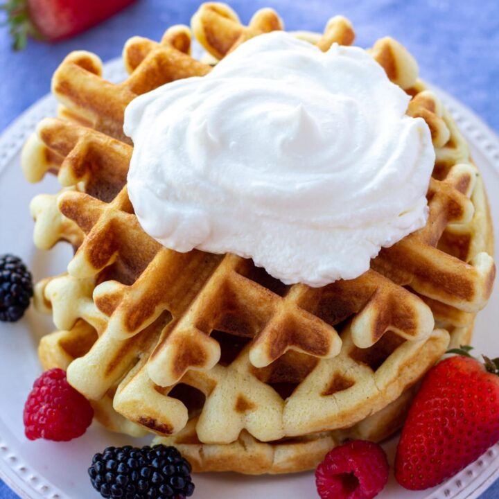 a stack of waffles with whipped cream, on a white plate, surrounded by berries