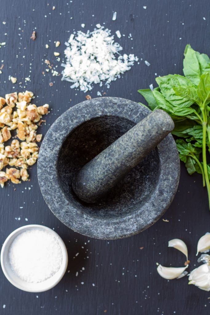 salt, parmesan, walnuts, basil, and garlic surrounding an empty mortar with a pestle in the middle