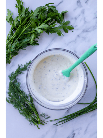 ranch dressing in a glass bowl with herbs surrounding it on a marble table