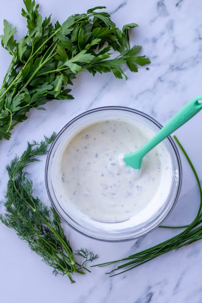 ranch dressing in a glass bowl with herbs surrounding it on a marble table