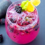 blackberry fizz in a clear glass with whole berries in and around the glass on a black background