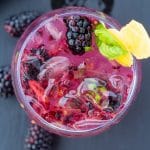 blackberry fizz in a clear glass with whole berries in and around the glass on a black background