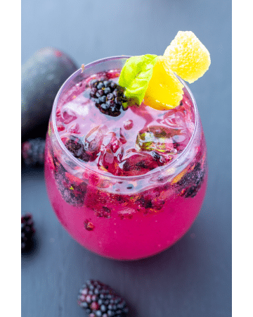 blackberry fizz in a clear glass with whole berries in and around the glass on a black background