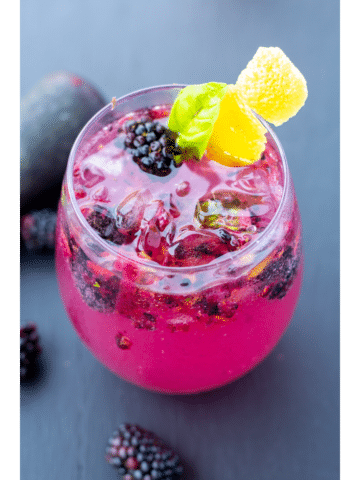 blackberry fizz in a clear glass with whole berries in and around the glass on a black background