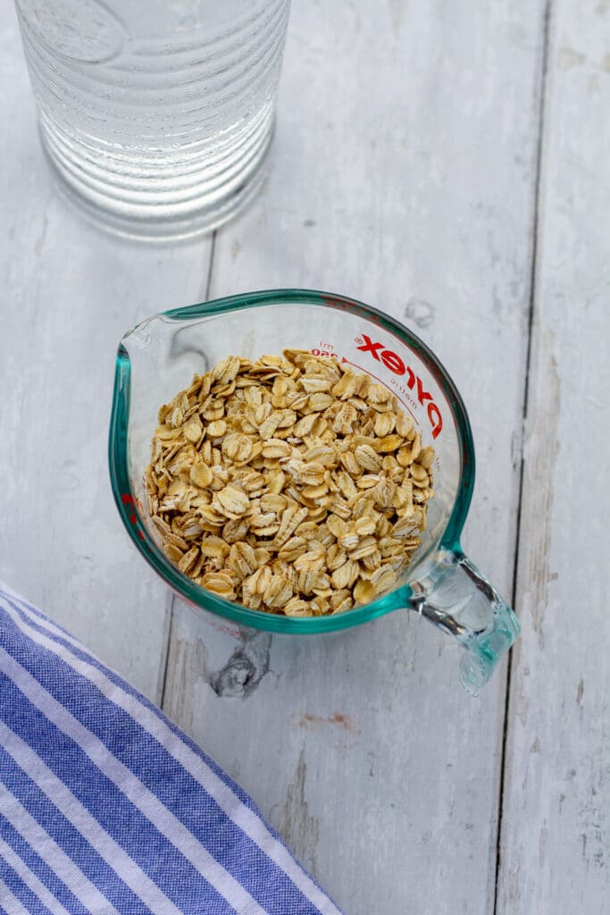 oats in a glass measuring cup next to a blue and white cloth and a jar of water on a white background