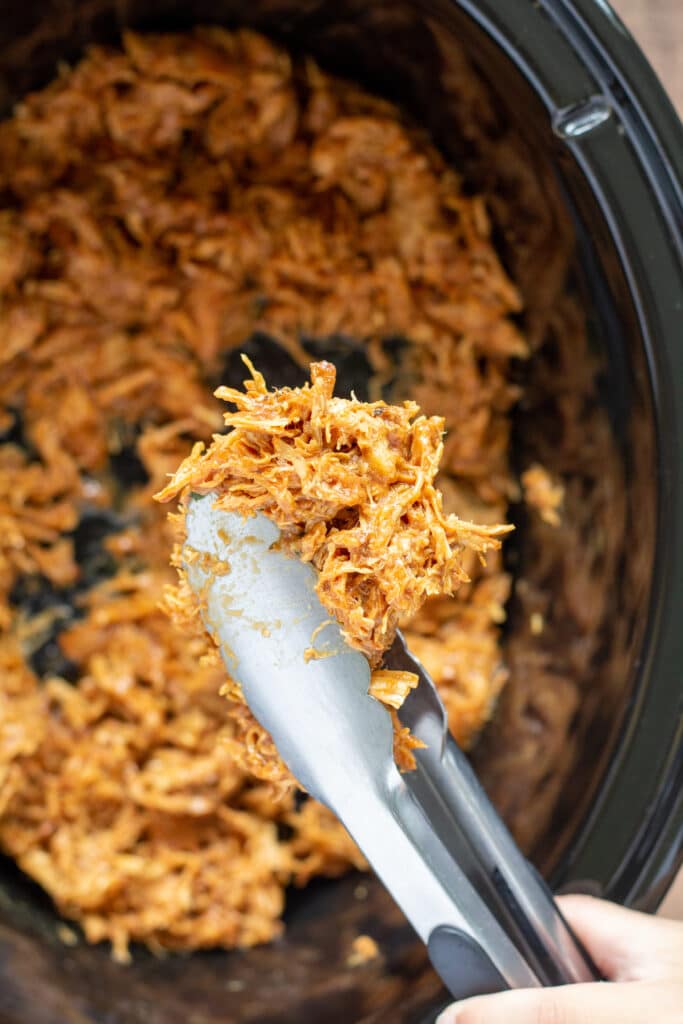 shredded chicken held by tongs over a crockpot of more chicken