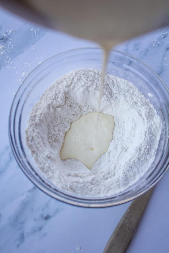 Cream pouring into a flour mixture in a glass bowl on a white marble counter