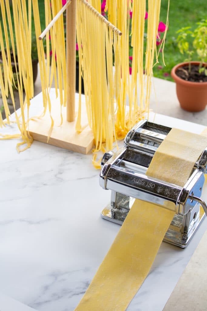 a piece of dough in a pasta machine next to a rack of cut pasta