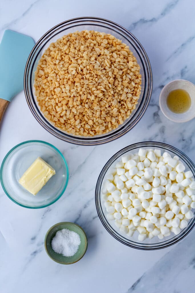 Rice cripies, marshmallows, butter, salt, and vanilla in bowls on a white marble surface