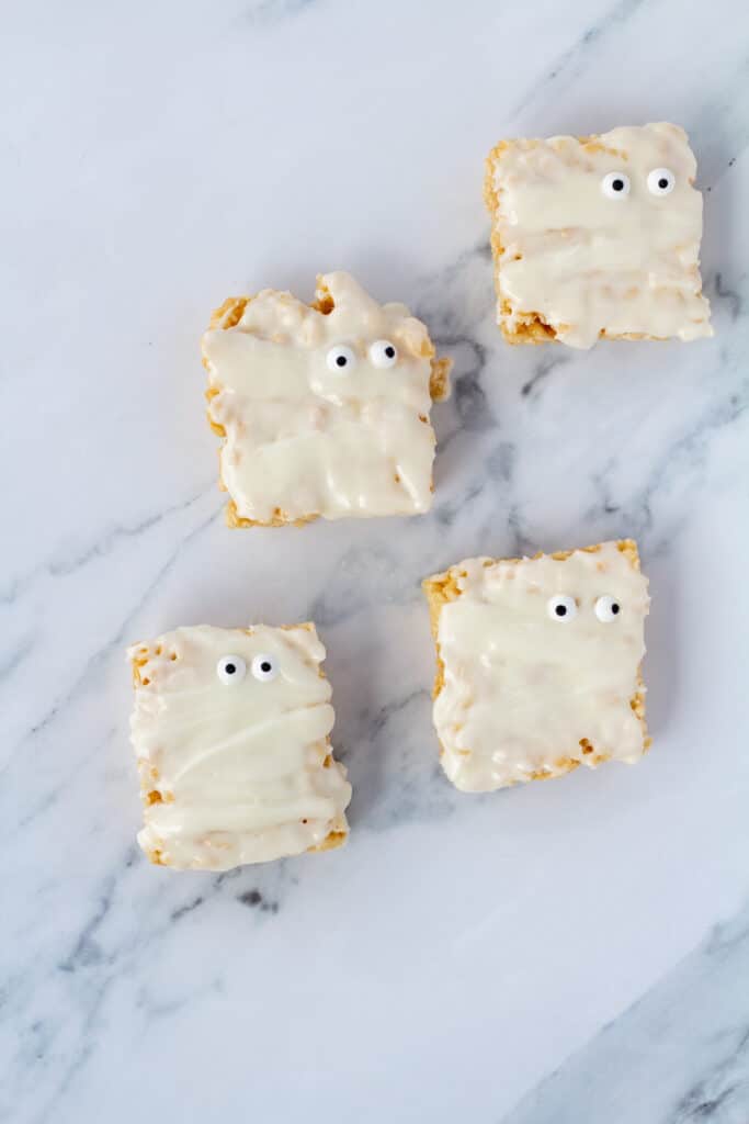 Four mummy rice crispy treats covered with icing with googly eyes on a white marble counter