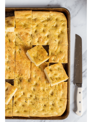 sliced focaccia bread in a sheet pan on a marble table with a long serrated knife next to the pan