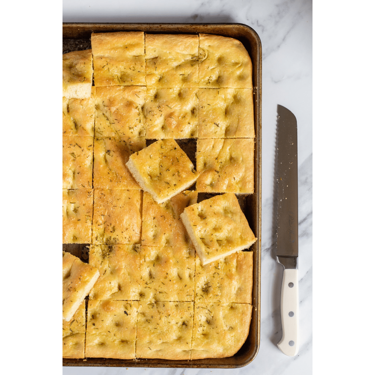 sliced focaccia bread in a sheet pan on a marble table with a long serrated knife next to the pan