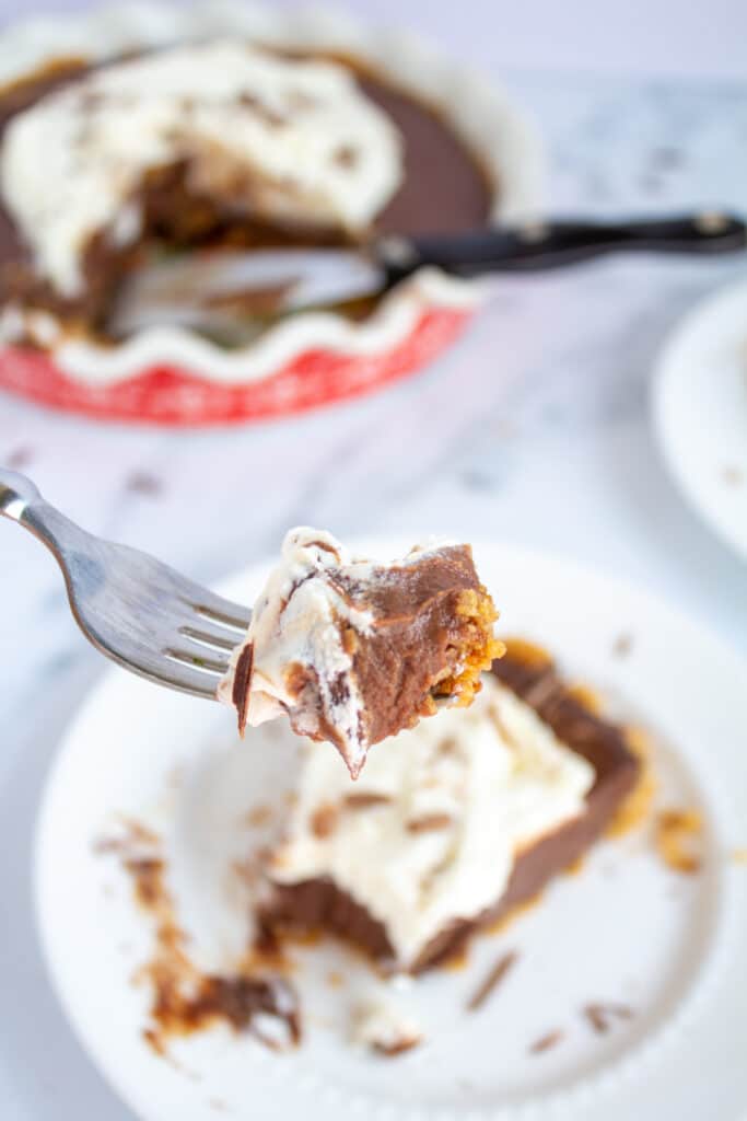 a close up of a fork with a piece of chocolate pie and whipped cream with the slice and pie in the background