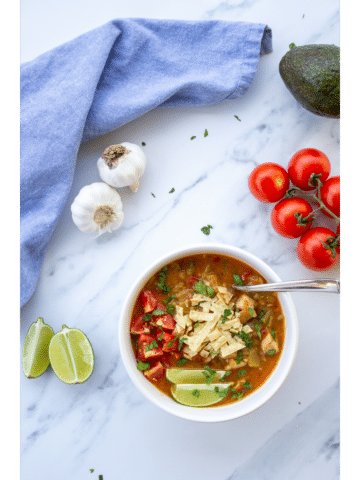 green chile in a bowl on a marble surface with tomatoes, garlic, limes, and avocado surrounding it