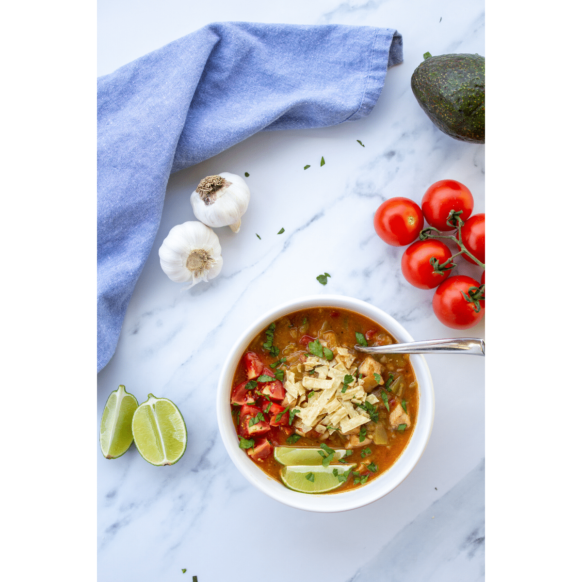 green chile in a bowl on a marble surface with tomatoes, garlic, limes, and avocado surrounding it