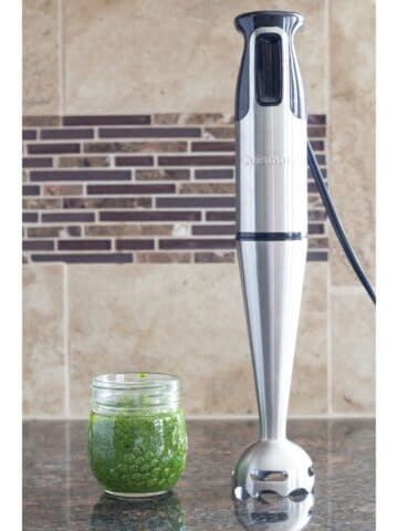 an immersion blender standing on a counter and sitting next to pesto