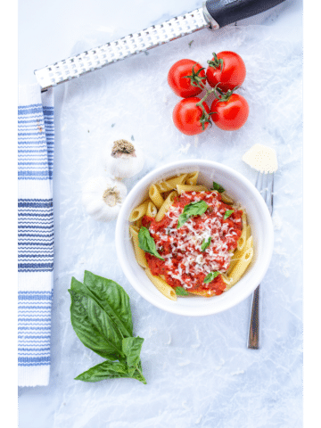 marinara sauce in a white bowl surrounded by garlic, basil leaves, tomatoes on the vine, a fork, and a white and blue napkin