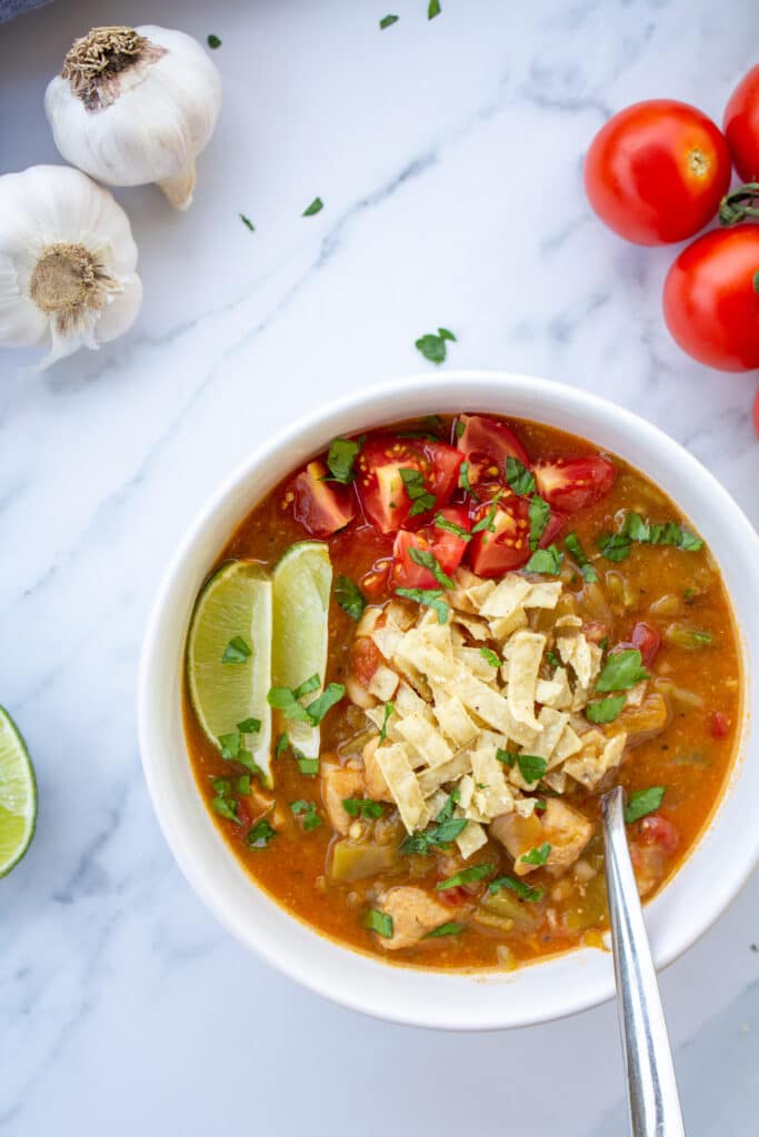 green chile in a bowl on a marble surface with tomatoes, garlic, limes, and avocado surrounding it