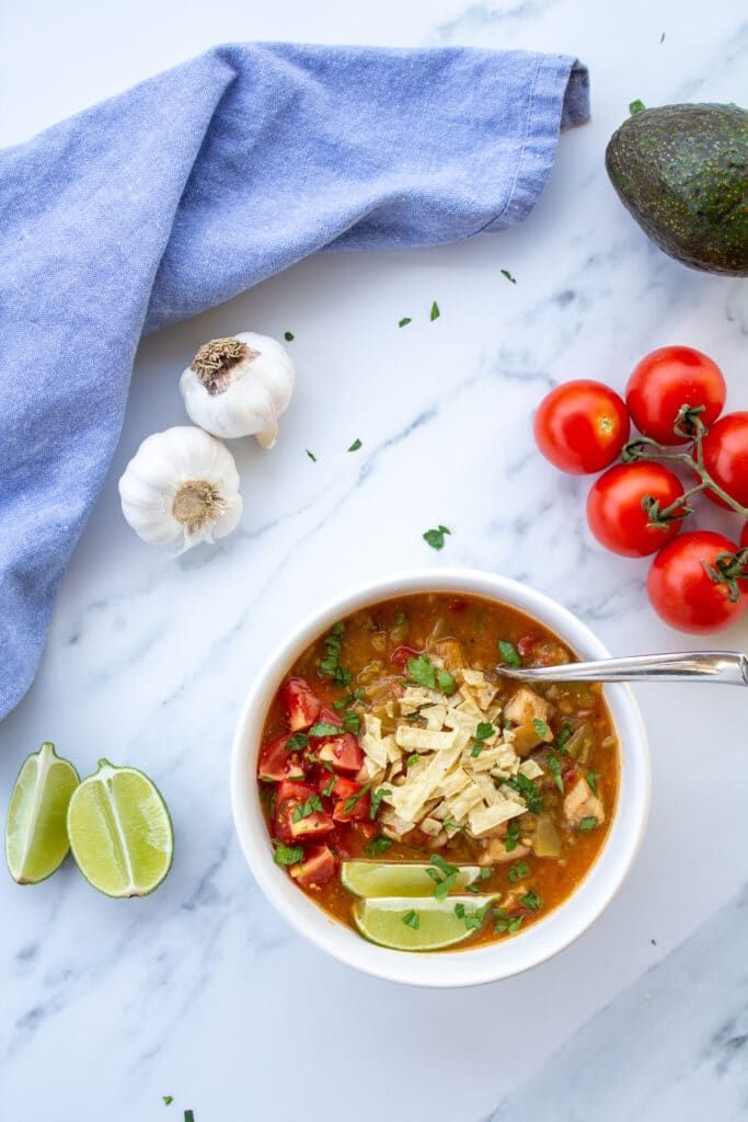 green chile in a bowl on a marble surface with tomatoes, garlic, limes, and avocado surrounding it