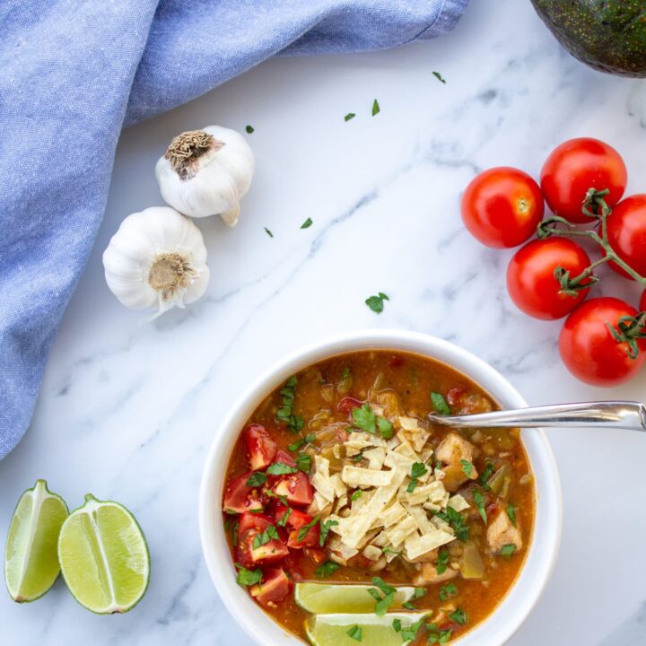 green chile in a bowl on a marble surface with tomatoes, garlic, limes, and avocado surrounding it