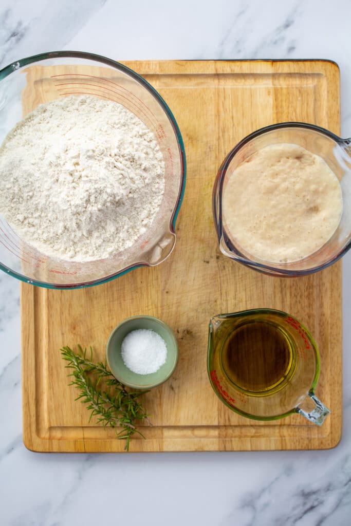 a bowl of flour, a bowl of yeast, a bowl of salt, a bowl of olive oil, a piece of rosemary all sitting on a cutting board