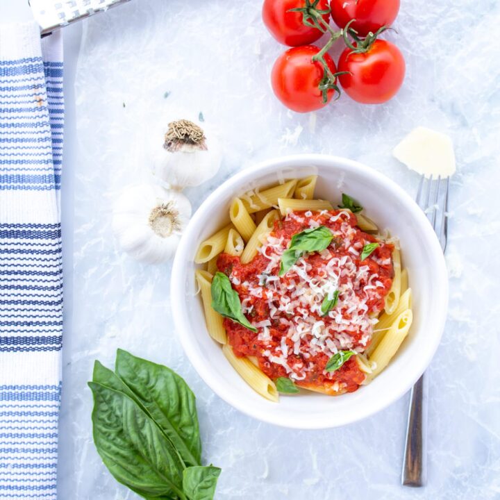 marinara sauce in a white bowl surrounded by garlic, basil leaves, tomatoes on the vine, a fork, and a white and blue napkin