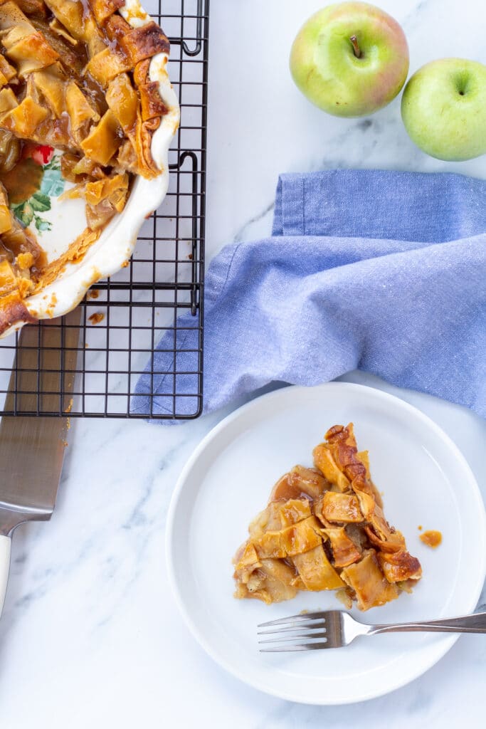 A slice of apple pie next to a cooling rack with a whole pie next to it