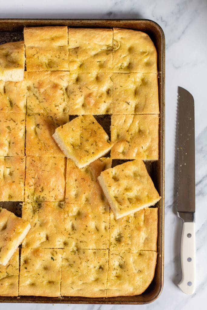 sliced focaccia bread in a sheet pan on a marble table with a long serrated knife next to the pan