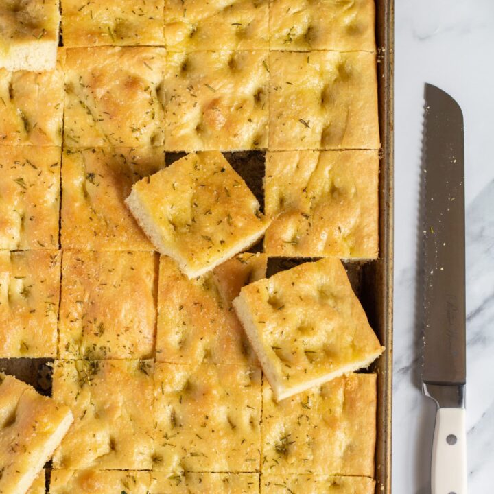 sliced focaccia bread in a sheet pan on a marble table with a long serrated knife next to the pan